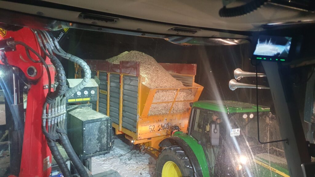 Nächtliche Produktion von Holzhackschnitzeln auf Gut Tangstedt: Ein Traktor mit beladenem Anhänger steht im Scheinwerferlicht neben einer Häckselmaschine im verschneiten Außenbereich.
