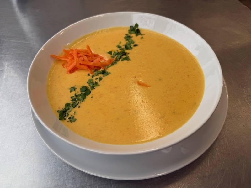 Cremige Karotten-Ingwer-Suppe in einer wei&szlig;en Schale, ansprechend garniert mit frischen Gem&uuml;seraspeln und einer Spur gehackter Petersilie.